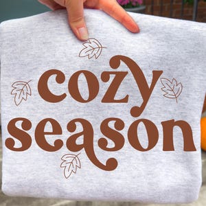 May include: A light grey heather sweatshirt featuring the brown text "cozy season" printed in a bold, rounded font. The design is accented with four simple, thin-lined brown leaf illustrations positioned around the text on the front of the garment.