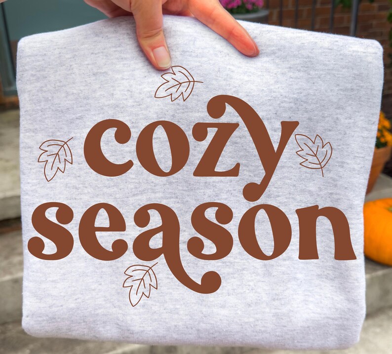 May include: A light grey heather sweatshirt featuring the brown text "cozy season" printed in a bold, rounded font. The design is accented with four simple, thin-lined brown leaf illustrations positioned around the text on the front of the garment.