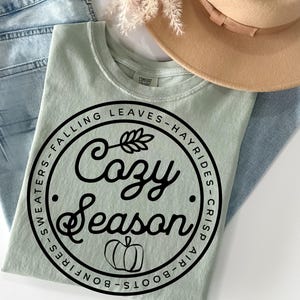 May include: A sage green t-shirt featuring a black circular graphic with the text "Cozy Season" in script, surrounded by the words "Falling Leaves", "Hayrides", "Crisp Air", "Boots", "Bonfires", and "Sweaters", with a small pumpkin icon.