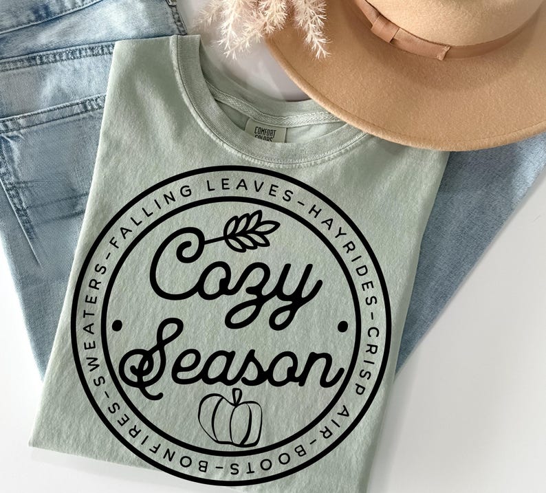 May include: A sage green t-shirt featuring a black circular graphic with the text "Cozy Season" in script, surrounded by the words "Falling Leaves", "Hayrides", "Crisp Air", "Boots", "Bonfires", and "Sweaters", with a small pumpkin icon.