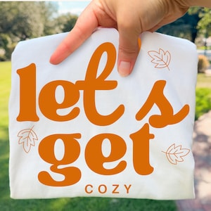 May include: A white t-shirt featuring the orange text "let's get" in a large, bold, rounded font, with the word "COZY" printed below in smaller, simple orange letters. The design is accented with small, thin-lined orange leaf illustrations.