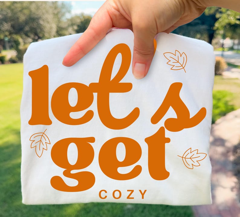 May include: A white t-shirt featuring the orange text "let's get" in a large, bold, rounded font, with the word "COZY" printed below in smaller, simple orange letters. The design is accented with small, thin-lined orange leaf illustrations.