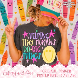 May include: A charcoal gray t-shirt featuring the text "HELPING TINY HUMANS DO BIG THINGS" in colorful, playful lettering, accompanied by pink stars and a large yellow smiley face. The shirt is paired with a multi-colored trucker hat.