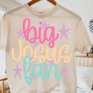 K&ouml;nnte beinhalten: Beiges T-Shirt mit den Worten "big Jesus fan" in rosa, gelber und t&uuml;rkisfarbener Schrift, mit lila Sternakzenten. Das T-Shirt h&auml;ngt an einem Holzb&uuml;gel.