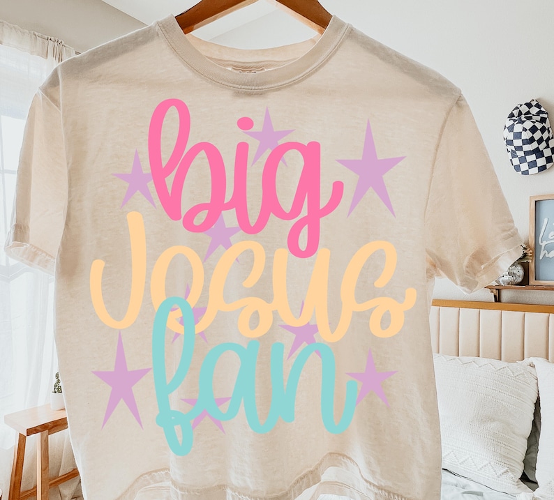 K&ouml;nnte beinhalten: Beiges T-Shirt mit den Worten "big Jesus fan" in rosa, gelber und t&uuml;rkisfarbener Schrift, mit lila Sternakzenten. Das T-Shirt h&auml;ngt an einem Holzb&uuml;gel.