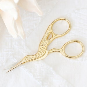May include: Gold-toned bird-shaped sewing scissors with intricate details on a white lace background.