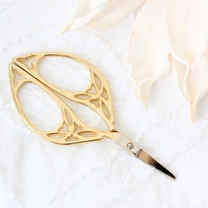 May include: Gold-toned decorative scissors with a floral design. The scissors have a small, pointed blade and a loop handle.