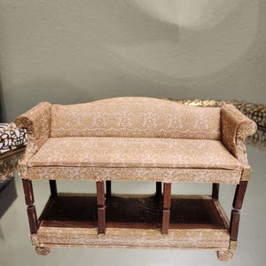May include: Miniature sofa with a beige and gold damask pattern. The sofa has dark brown wooden legs and a lower shelf. The arms are curved, and the back is slightly arched. A charming piece for a dollhouse or miniature collection.