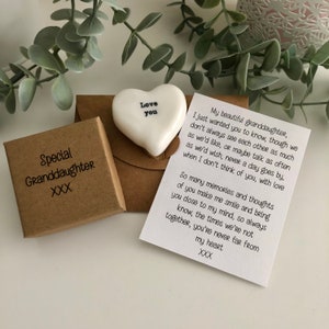 May include: A small white ceramic heart with the words "Love you" on it, sitting on a brown envelope with a handwritten note inside. The envelope is next to a small brown box with the words "Special Granddaughter XXX" on it.