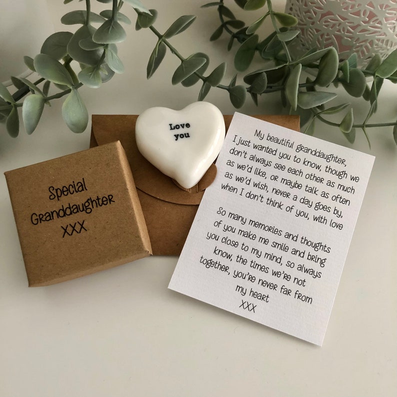 May include: A small white ceramic heart with the words "Love you" on it, sitting on a brown envelope with a handwritten note inside. The envelope is next to a small brown box with the words "Special Granddaughter XXX" on it.