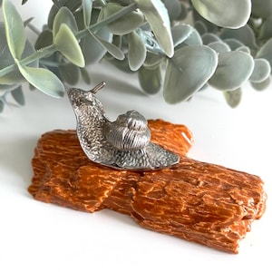 May include: A silver snail ornament sits on a brown, textured, log-shaped base. The snail has detailed shell and body features. The background includes green foliage, creating a naturalistic display. The ornament is likely a decorative item.
