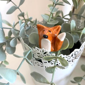 May include: A small ceramic fox figurine with orange and white paint, sitting in a white planter with green eucalyptus leaves.