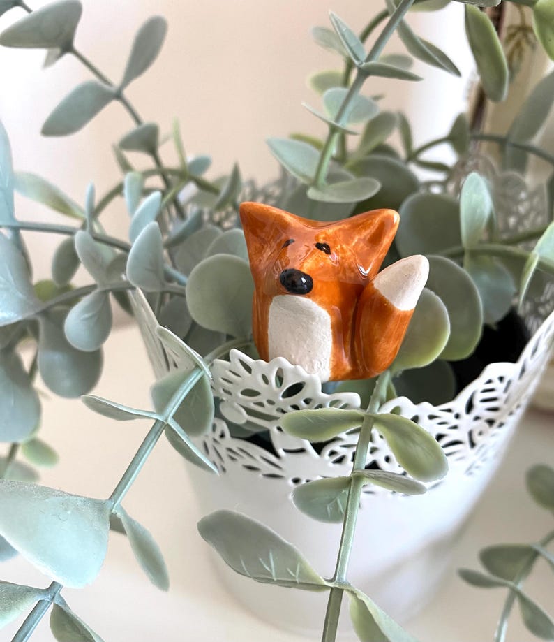 May include: A small ceramic fox figurine with orange and white paint, sitting in a white planter with green eucalyptus leaves.