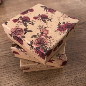 May include: Three square boxes stacked on top of each other. The boxes are made of brown cardboard and have a floral pattern with red roses.