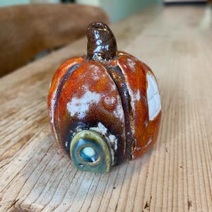 May include: A ceramic pumpkin figurine with a glossy orange and brown glaze, featuring a dark brown stem, a small arched doorway with a blue accent, and a white window detail on the side, crafted with a rounded shape and textured surface finish.