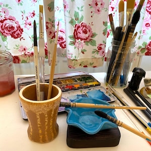 May include: An assortment of art supplies, including paintbrushes, watercolour paints, and a small blue dish. The paintbrushes have wooden handles and black bristles. The watercolour paints are in a metal palette.