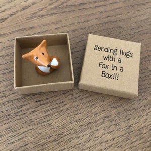 May include: A small ceramic fox figurine holding a heart in its paw, inside a brown cardboard box. The box has the text "Sending Hugs with a Fox in a Box!!!" printed on it.