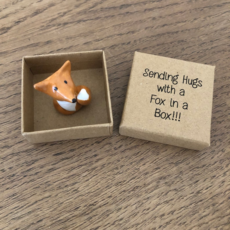 May include: A small ceramic fox figurine holding a heart in its paw, inside a brown cardboard box. The box has the text "Sending Hugs with a Fox in a Box!!!" printed on it.