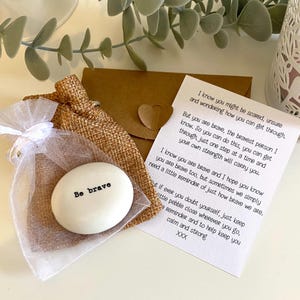 May include: A white, oval stone inscribed with "Be brave" in black, displayed in a sheer white organza bag and a burlap pouch. A small card with a heart-shaped closure and a handwritten message is also included.