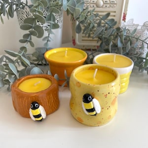 May include: Four handmade candles in ceramic containers. The candles are filled with yellow wax and have white wicks. One candle is in a tree stump-shaped container with a bee. Other containers are yellow, orange, and white.