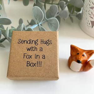 May include: A small brown box with the text "Sending Hugs with a Fox in a Box!!!" printed on the top. A small ceramic fox figurine is sitting next to the box.