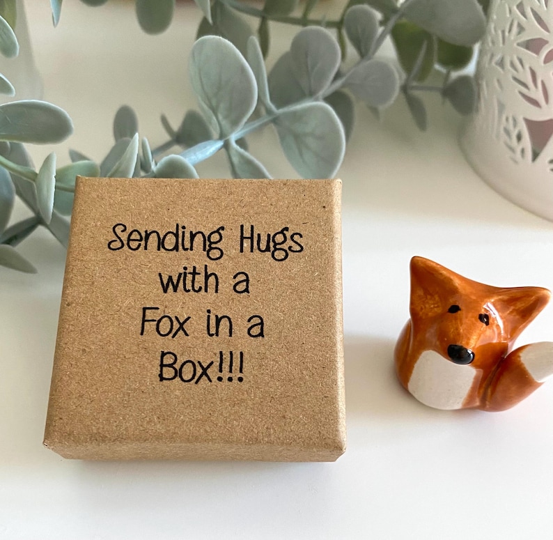 May include: A small brown box with the text "Sending Hugs with a Fox in a Box!!!" printed on the top. A small ceramic fox figurine is sitting next to the box.