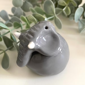 May include: A grey ceramic elephant ornament with white tusks and a trunk. The elephant has two holes on top, suggesting it is a salt or pepper shaker. The ornament is on a white surface with green leafy sprigs in the background.