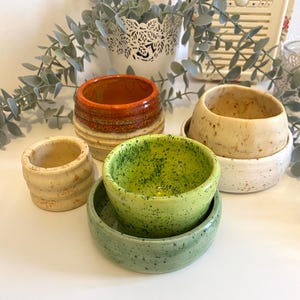 May include: A selection of small ceramic bowls in different sizes and colours. The bowls have speckled and textured finishes in shades of green, orange, and cream. Some bowls are stacked, showing their different shapes and designs.