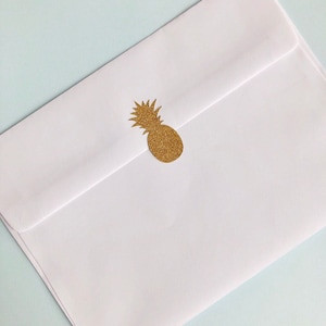 Può includere: Una busta bianca con un adesivo a forma di ananas con glitter dorati sul davanti.