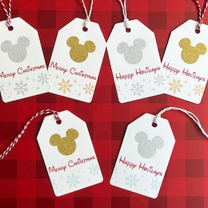 Puede incluir: Etiquetas de regalo blancas con un diseño de cabeza de Mickey Mouse en purpurina plateada y dorada. Las etiquetas tienen hilo rojo y blanco y dicen "Merry Christmas" y "Happy Holidays" con acentos de copo de nieve.