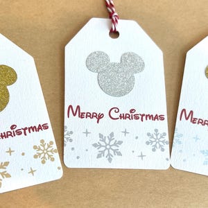 May include: Three white gift tags with a Mickey Mouse head design in gold and silver glitter. The tags have the words "Merry Christmas" in red script and snowflake accents. Each tag has a string for hanging.