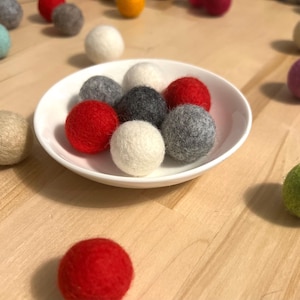 May include: A white bowl filled with colorful felt balls. The balls are red, white, and gray. The bowl is sitting on a wooden surface.