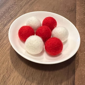 May include: A white ceramic bowl filled with seven red and white felt balls. The balls are arranged in a circular pattern, with the red balls alternating with the white ones. The bowl is on a wooden surface.