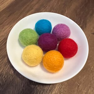 May include: A white ceramic bowl filled with seven colorful felt balls. The balls are in shades of blue, green, purple, yellow, orange, and red. The felt balls are round and have a textured appearance.
