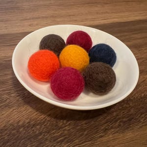 May include: A white, oval-shaped bowl filled with seven colorful felt balls. The balls are in shades of orange, yellow, burgundy, dark brown, and navy blue. The bowl sits on a dark brown wooden surface.