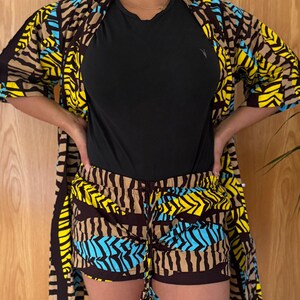 May include: A matching set of clothing featuring a black top, shorts, and a kimono-style jacket. The shorts and jacket have a vibrant pattern with brown, yellow, and blue geometric shapes. The outfit is worn by a person.