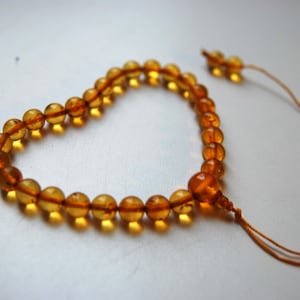 Könnte beinhalten: Ein Armband aus durchscheinenden, honigfarbenen Bernsteinperlen, die in Herzform angeordnet sind. Die Perlen sind auf eine dünne, hellbraune Schnur gefädelt, die an den Enden herausragt, mit einer größeren Mittelperle und einem kleinen Knoten.