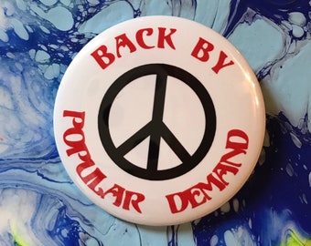 Könnte beinhalten: Ein weißer Knopf mit einem schwarzen Friedenssymbol und dem Text "Back by Popular Demand" in roten Buchstaben. Der Knopf befindet sich auf einem bunten Hintergrund aus Blau, Gelb und Weiß.