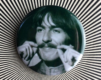 May include: A black and white photo of a man with a mustache. He is holding his mustache with his fingers. The photo is on a green button.