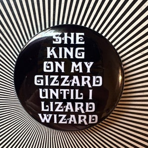 May include: A black button with white text that reads "She King On My Gizzard Until I Lizard Wizard".