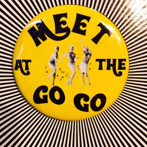 Puede incluir: Botón amarillo con texto negro que dice "Meet at the Go Go". Tres mujeres en blanco y negro están bailando en el centro del botón.