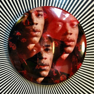 May include: A red, white, and black button featuring a close-up portrait of a person with a floral pattern in the background.
