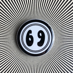May include: A black and white button with the number 69 in a white circle on a black background. The button is on a black and white striped background.