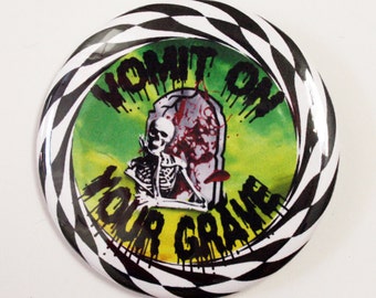 May include: A round button with a black and white checkered border. The center features a green and yellow background with a skeleton, tombstone, and the words "Vomit on Your Grave" in black lettering. The image has a horror theme.