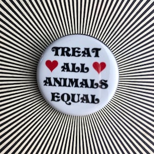 Könnte beinhalten: Weißer Knopf mit schwarzem Text, der "Treat All Animals Equal" lautet, mit zwei roten Herzen auf beiden Seiten des Wortes "All".
