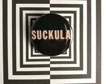 May include: A black button with the word "SUCKULA" printed in white letters.
