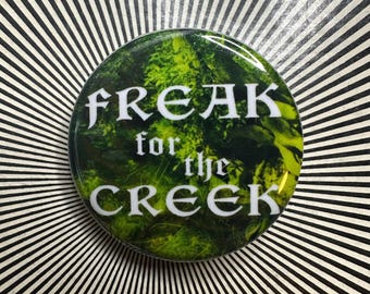 May include: A circular badge with the words "FREAK for the CREEK" in white, decorative lettering. The badge features a green and yellow forest scene. The background is a black and white striped pattern.