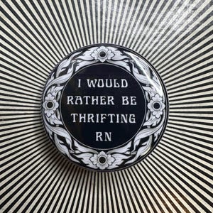 Puede incluir: Un botón redondo en blanco y negro con el texto "I WOULD RATHER BE THRIFTING RN" en blanco. El botón tiene un diseño floral y está sobre un fondo a rayas blancas y negras.
