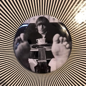 Puede incluir: Botón de foto en blanco y negro con un hombre sentado con los pies en una guitarra, sosteniendo una taza de té. El hombre tiene el pelo corto y oscuro y lleva una camisa de color claro.