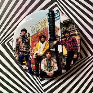 Puede incluir: Una foto vintage de cuatro hombres con ropa de los años 60 de pie frente a una escena de calle de la ciudad con letreros y anuncios. Los hombres llevan camisas estampadas, jeans y bufandas. La foto está en un marco redondo.